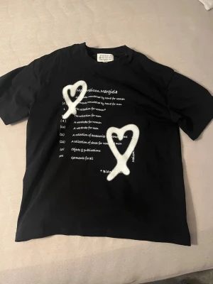 Svart Maison Margiela t-shirt med hjärtan - Cool svart t-shirt från Maison Margiela med vit text och två stora sprayade hjärtan på framsidan. Klassisk passform och rund hals. Perfekt för dig som gillar streetwear och vill sticka ut med en unik design.