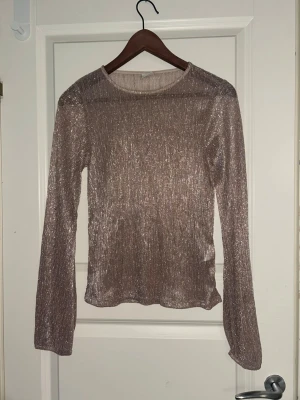 Glittrig långärmad topp i rosa - Säljer en glittrig rosa långärmad topp med rund hals. Materialet är stretchigt och skimrar snyggt i ljuset, perfekt för att sticka ut. Passformen är normal och toppen är tunn och lätt, vilket gör den enkel att styla till olika looks. Färgen på sista bilden är färgen på tröjan. Tröjan är relativt genomskinlig ovksp