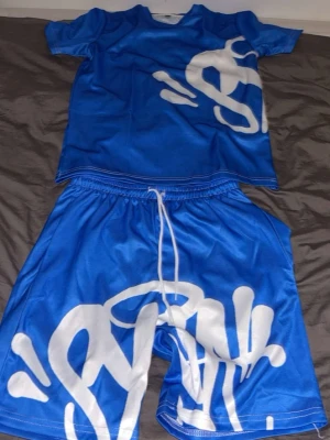 Blå shorts med vit graffiti - Säljer ett par blå shorts med vit graffiti-inspirerad print och snörning i midjan. Shortsen har en loose passform och är gjorda i ett glansigt syntetmaterial som känns sportigt och bekvämt. Perfekta för dig som vill sticka ut med en streetwear-look.