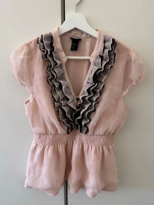 Blus - Supersöt ljusrosa blus med djup v-ringning och dubbla volanger och svart spets längs urringningen😋. Den är tillverkad i ett tunt, skirt tyg. Säljs pgv att den är för stor🙃