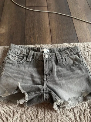 Grå jeansshorts från Gina Tricot - Säljer ett par grå jeansshorts från Gina Tricot i storlek 158. Shortsen har råa, fransiga kanter och klassisk femficksdesign med bälteshällor. Perfekta för varma dagar och ger en avslappnad look.