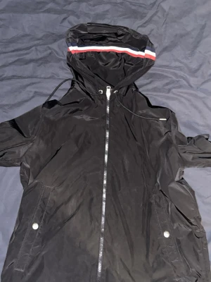 Svart vindjacka med huva från Moncler - Snygg svart vindjacka från Moncler med dragkedja framtill, två fickor med tryckknappar och justerbar huva. Huvan har en röd, vit och blå rand som detalj. Jackan är tillverkad i ett tunt, vindtätt material och har Moncler-logga på bröstet. Pris kan diskuteras vid snabb affär!!