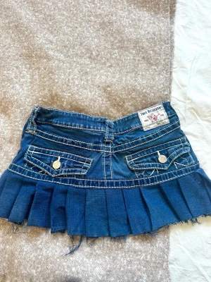 Blå jeanskjol - Säljer en blå jeanskjol från True Religion med plisserad kant och råa kanter. Kjolen har dekorativa fickor med knappar bak och klassiska jeansdetaljer framtill. 