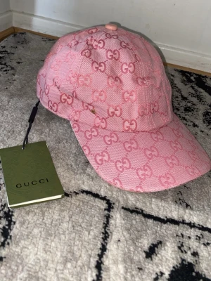 Rosa keps från Gucci med GG-mönster - Säljer en snygg rosa keps från Gucci med det klassiska GG-monogrammet över hela kepsen. Kepsen har böjd skärm och justerbar rem bak. Materialet är kraftigt tyg som ger en exklusiv känsla. Perfekt för dig som vill sticka ut med en trendig accessoar.