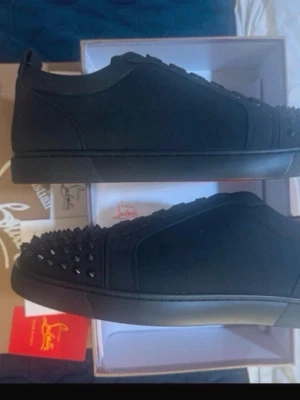 Bror kan du lägga mina loubs  - Snygga svarta sneakers från Christian Louboutin med mattsvarta nitar på tån och klassisk röd sula. Skorna har en låg profil, snörning och är tillverkade i mockaliknande material. Perfekta för dig som vill sticka ut med en edgy och lyxig stil.