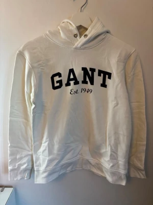 Vit hoodie från GANT  - Snygg vit hoodie från GANT med svart tryck på bröstet där det står 'GANT Est. 1949'. Hoodien har huva med metallöljetter och långa ärmar. Tillverkad i mjukt material som känns skönt mot huden. Perfekt för en avslappnad och trendig stil.