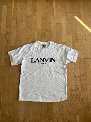 Vit Lanvin Paris t-shirt - Säljer en klassisk vit t-shirt från Lanvin Paris med svart tryck framtill. T-shirten har korta ärmar, rund halsringning och är tillverkad i mjuk bomull. Perfekt för en clean och stilren look.