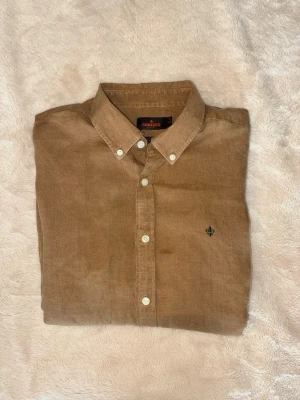 Beige Morris linneskjorta - Snygg beige Morris skjorta i linne med klassisk krage och vita knappar. Skjortan har lång ärm och en liten broderad detalj på bröstet. Perfekt för dig som gillar en avslappnad men stilren look.