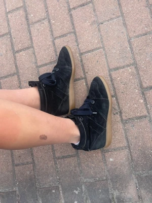 Isabel Marat skor med kilklack - Säljer ett par svarta sneakers i mocka med kilklack och gummisula från Isabel marant. Skorna har svarta skosnören och dekorativa sömmar. Perfekta för dig som vill ha en sportig men ändå trendig look. Bläddra för att se små defekter. 