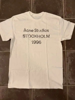 Vit Acne Studios t-shirt Stockholm - Vit t-shirt från Acne Studios med tryck i svart text på bröstet där det står 'Acne Studios STOCKHOLM 1996'. Klassisk rund hals och korta ärmar. Perfekt för en clean och stilren look. Acne är lite oversize men den sitter som en vanlig S