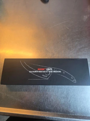 Huusk japansk kockkniv i presentförpackning - Säljer en Huusk japansk kockkniv i svart presentförpackning. Kniven har ett handtag i trä och ett blad i stål med hamrad yta och ett hål för bättre grepp. Perfekt för matlagning och snygg att ha i köket. Aldrig använd!