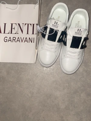 Valentino Rockstud Untitled sneakers - Säljer ett par vita Valentino Rockstud Untitled sneakers med svart band och nitar över vristen. Skorna har klassisk låg profil, vita snören och är tillverkade i slätt läder. Snygg och stilren design som sticker ut med den svarta detaljen.