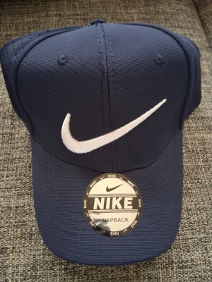 Mörkblå Nike keps med vit logga - Snygg mörkblå snapback-keps från Nike med den klassiska vita swoosh-loggan framtill. Kepsen har böjd skärm och ventilerande mesh på sidorna för extra komfort. Justerbar baktill för perfekt passform. Perfekt för en sportig och avslappnad stil.
