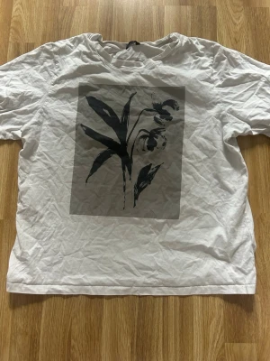 Vit t-shirt med svart blommotiv - Vit t-shirt med ett stort svart blommotiv på framsidan. Klassisk passform och rund halsringning. Perfekt för dig som gillar stilrena tryck och enkel design. Köpte den för 500 kr på Lindex, därför är priset lite dyrare än vanligt. Priser kan diskuteras.