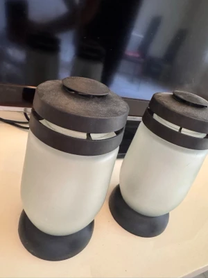 Två frostade ljuslyktor i glas - Säljer två stilrena ljuslyktor i frostat glas med svarta metallock och fot. Perfekta för värmeljus eller små blockljus. Modern design som passar i de flesta rum och ger ett mysigt sken.