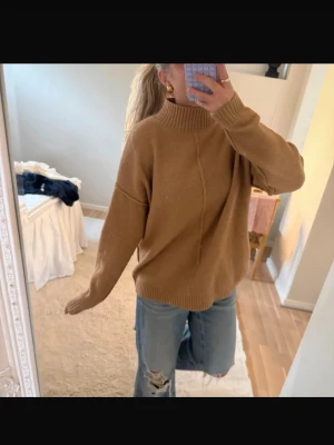 Beige oversized stickad tröja - Säljer en beige  stickad tröja med hög krage och ribbade muddar. Tröjan har en avslappnad passform och en dekorativ söm framtill. Perfekt att styla med jeans för en chill look.