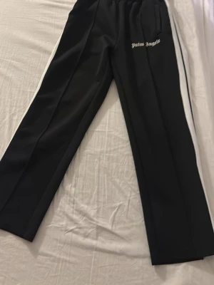 Palm Angels mjukis byxor - Säljer ett par svarta mjukisbyxor från Palm Angels med vita sidoränder och logga på vänster sida. Byxorna har rak passform, resår i midjan och sidofickor. Perfekta för en avslappnad och trendig streetwear-look.