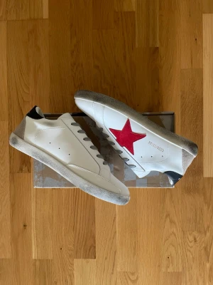 Vita sneakers från Golden Goose - Snygga vita sneakers från Golden Goose med en stor röd stjärna på sidan och grå detaljer på häl och sula. Skorna har svarta detaljer vid hälen och grå snören. Tillverkade i läder med en klassisk låg siluett som passar till det mesta.
