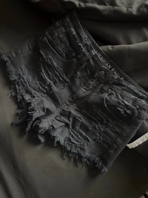 Svarta slitna shorts från American Eagle - Säljer ett par svarta, slitna shorts från American Eagle med råa kanter och flera revor för en edgy look. Gjorda i mjuk bomull med lite stretch för extra komfort. Perfekta för dig som gillar en avslappnad och trendig stil. Mer i storleks guiden finns på bild nr 2 har inte använt dom då dom ej passar. Tycker dock inte att dom är så stretchiga