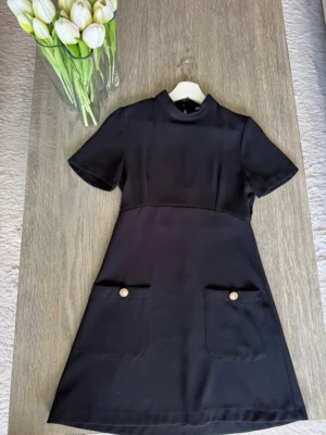 Svart kortklänning med fickor Zara - Stilren svart kortklänning med hög krage och korta ärmar. Klänningen har två stora fickor framtill, dekorerade med guldknappar. Rak passform och enkel design som ger en klassisk look. Fint skick 