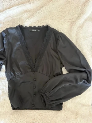 Svart Satin topp Bikbok - Jättefin svart topp från BikBok i ett elegant satinmaterial med fina, lätt ballongformade ärmar. Framtill har den en djup V-ringning med dekorativ spets som ger toppen en feminin och lite lyxigare känsla. Baksidan är stretchig och har ett smockat parti med spetsdetalj, vilket gör att den sitter fint och följer kroppen. • Storlek XS • Satinmaterial • Lätt ballongärmar • V-ringad med spetsdetalj • Stretchig/smockad rygg • Använd fåtal gånger • Inga defekter • Mycket fint skick 🤍