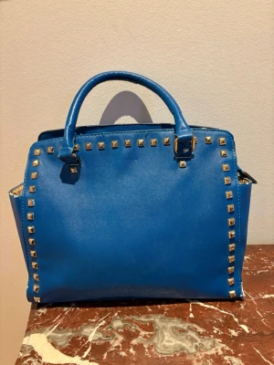 Blå handväska med nitar - Välj gärna instabox. Kan skicka snabbare då. Snygg blå handväska från Michael Kors i slätt läder med guldfärgade nitar runt kanterna. Väskan har två handtag och ett rymligt innerfack med flera mindre fack och dragkedja. Insidan är beige med MK-logga. Perfekt för dig som vill sticka ut.
