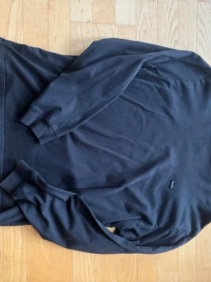 Svart långärmad tröja från Supreme - Svart långärmad tröja från Supreme med diskret logotyp på sidan. Tillverkad i mjukt bomullsmaterial och har ribbade muddar vid ärmsluten. Enkel och clean design som passar perfekt till streetwear-stilen.