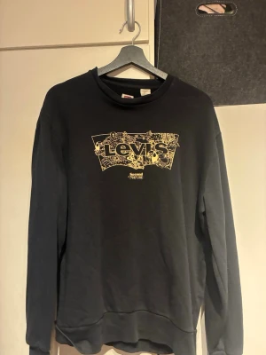 Svart sweatshirt från Levi's M - Cool svart sweatshirt från Levi's med ett stort gult logotryck på bröstet och detaljerat mönster i trycket. Tröjan har rund hals, långa ärmar och ribbade muddar. Perfekt för en avslappnad och trendig stil.