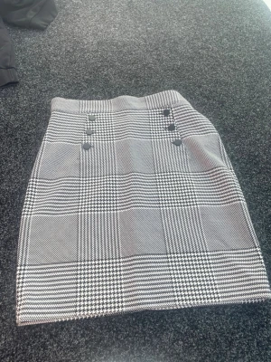 Rutig kortkjol med knappar - Snygg rutig kortkjol i svart och vitt med dekorativa knappar framtill och dragkedja i sidan. Kjolen har en klassisk rak passform och är perfekt för att skapa en trendig look. Materialet känns mjukt och har en lätt struktur.