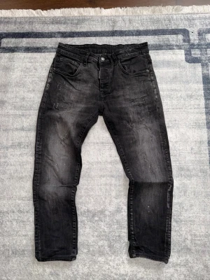 Svarta slim fit jeans med slitningar - Säljer ett par svarta slim fit jeans med coola slitningar och lätt tvättad look. Jeansen har fem fickor, dragkedjegylf och detaljer som ger en trendig streetkänsla. Perfekta för dig som gillar en avslappnad men ändå snygg stil.