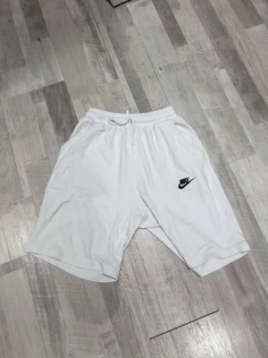 Vita Nike shorts med snörning - Säljer ett par vita shorts från Nike med elastisk midja och snörning för skön passform. Shortsen har en klassisk Nike-logga på ena benet och är tillverkade i mjukt material, perfekta för sommardagar eller träning.