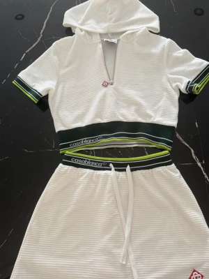 Vitt ribbat set från Casablanca - Snyggt vitt ribbat set från Casablanca med croppad topp med huva och dragkedja . Kjolen har resår och dragsko i midjan samt inbyggda shorts för extra komfort.