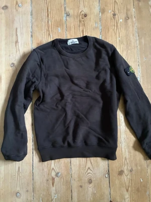 Svart sweatshirt från Stone Island - Svart sweatshirt från Stone Island Junior med rund halsringning och klassisk logotyp-patch på vänster ärm. Tröjan har ribbade muddar vid ärmslut och nederkant, perfekt för en avslappnad och stilren look.