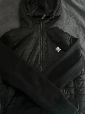 J Lindberg cardigan - Snygg svart cardiganjacka med quiltad front och stickade ärmar. Jackan har dragkedja, huva och en liten logotyp på bröstet. Perfekt för lager-på-lager den är är fin skick storlek S om intresse skriv elle mer bilder