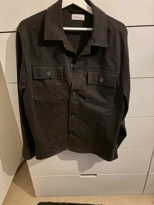Svart overshirt  - Stilren svart overshirt med klassisk krage och två stora bröstfickor med knappar. Skjortan har raka ärmar och knäppning framtill. Materialet ser ut att vara bomull eller en bomullsblandning, vilket ger en mjuk och bekväm känsla.
