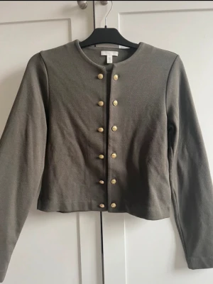Olivgrön kofta med guldfärgade knappar - Stilren olivgrön kofta med rund halsringning och dekorativa guldfärgade knappar framtill. Koftan har lång ärm och rak passform, perfekt att matcha med jeans eller kjol. Materialet känns mjukt och bekvämt mot huden.