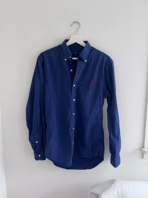 Blå skjorta med röd logga - Ralph Lauren - Klassisk blå skjorta med knappar framtill och button-down krage. Skjortan har lång ärm och en röd broderad logga på bröstet. Tillverkad i bomull och passar perfekt till en avslappnad men ändå stilren look.