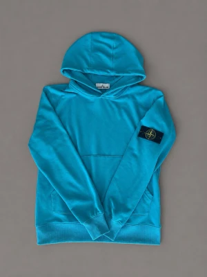 Turkos hoodie från Stone Island M - Snygg turkos hoodie från Stone Island med klassisk patch på vänster ärm. Hoodien har magficka framtill och ribbade muddar vid ärmslut och nederkant. Perfekt för dig som vill ha en streetwear-look med premiumkänsla. Materialet är mjukt och skönt bomullstyg.