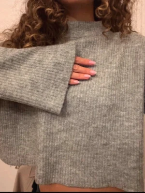 Grå ribbstickad tröja  - Mysig grå ribbstickad tröja med croppad passform och vida ärmar. Tröjan har hög hals och är tillverkad i ett mjukt stickat material. Perfekt för lager på lager och en chill look. (Finns noppror på tröjan)