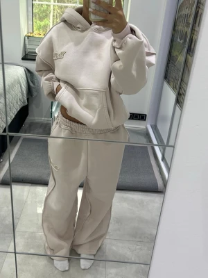 Beige oversized loungewear-set - Mysig och oversized loungewear-set i beige med hoodie och vida byxor. Hoodien har magficka och broderad detalj på bröstet, byxorna har resår i midjan och matchande broderi. Perfekt för chill dagar hemma.