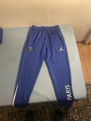 Blå PSG x Jordan träningsbyxor - Säljer ett par blå träningsbyxor från PSG x Jordan med vita detaljer. Byxorna har elastisk midja, PSG-logga på ena benet och Jumpman-logga på andra samt texten PARIS längs benet. Perfekta för fotboll eller chill.