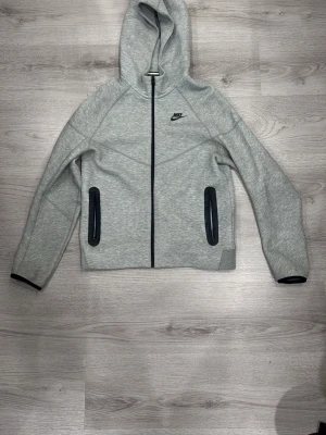 Grå zip hoodie Nike Tech - Snygg grå hoodie från Nike med dragkedja framtill och svarta detaljer vid fickorna. Klassisk Nike-logga på bröstet och huva för extra stilpoäng. Perfekt till chill eller streetwear-look. Tillverkad i mjukt material för skön känsla.