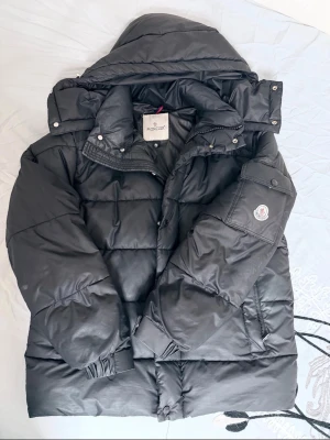 Svart dunjacka från Moncler - Snygg svart dunjacka från Moncler med huva och quiltad design. Jackan har dragkedja, tryckknappar och en Moncler-logga på ärmen. Perfekt för kalla vinterdagar och riktigt varm tack vare dunfyllningen.
