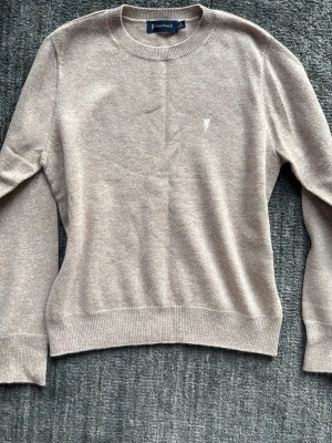 Beige stickad tröja från Raulp Lauren - Snygg beige stickad tröja från Raulp Lauren med rund halsringning och diskret broderad logga på bröstet. Tröjan har långa ärmar och ribbade muddar vid ärmslut och nederkant. Perfekt för lager på lager och stilrena outfits.