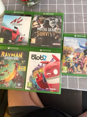 Xbox One spelpaket med 5 titlar - Säljer ett paket med fem Xbox One-spel: Assetto Corsa, Metal Gear Survive, World to the West, Rayman Legends och de Blob 2. Alla spel kommer i sina gröna plastfodral med färgglada omslag och varierande genrer som racing, action, äventyr och plattform.