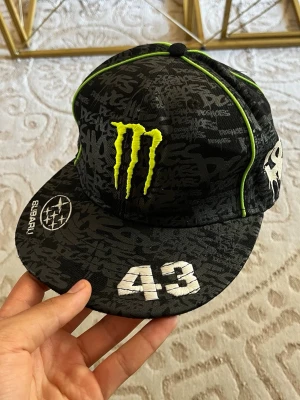 Monster x DC x Subaru svart keps - Säljer en svart keps med broderade loggor från Monster, DC och Subaru samt siffran 43. Kepsen har grön piping och grafiskt mönster över hela ytan. Platt skärm och justerbar baksida. Perfekt för dig som gillar streetwear och motorsport.