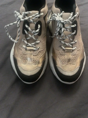 Chunky beige och svarta steve madden skor  - Chunky sneakers i beige, svart och guld med vit tjock sula. Skorna har meshdetaljer för extra ventilation och svarta/vita mönstrade skosnören. Snygg kontrast mellan färgerna och sportig design som sticker ut.