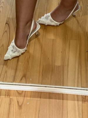 Vita spetsiga pumps med spets och blommor - Säljer ett par vita pumps med spetsig tå, dekorerade med spets och små 3D-blommor. De är i storlek 40 men jag har storlek 38-39 och de passar jättebra. Bandet runt hälen är elastiskt.
