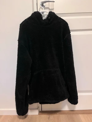 Svart teddyjacka med huva - En svart teddyjacka i supermjukt och fluffigt material. Jackan har huva, långa ärmar och en stor magficka framtill. Perfekt för dig som gillar en chill och cozy vibe. Passar till både jeans och mjukisbyxor.