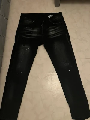 Dsquared 2 - Säljer ett par svarta slim fit jeans med coola slitningar och diskreta vita färgstänk. Jeansen har fem fickor, knappgylf och en tvättad look som ger en trendig vibe. Perfekta för dig som gillar streetstyle och vill ha något unikt i garderoben.